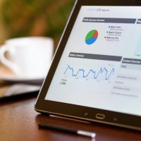 Close-up of a tablet displaying analytics charts on a wooden office desk, alongside a smartphone and coffee cup.